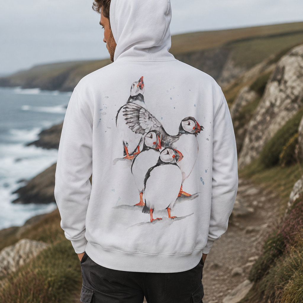 A man viewed from behind wearing a white hoodie with a large puffin illustration on the back, standing on a rocky coastal path.